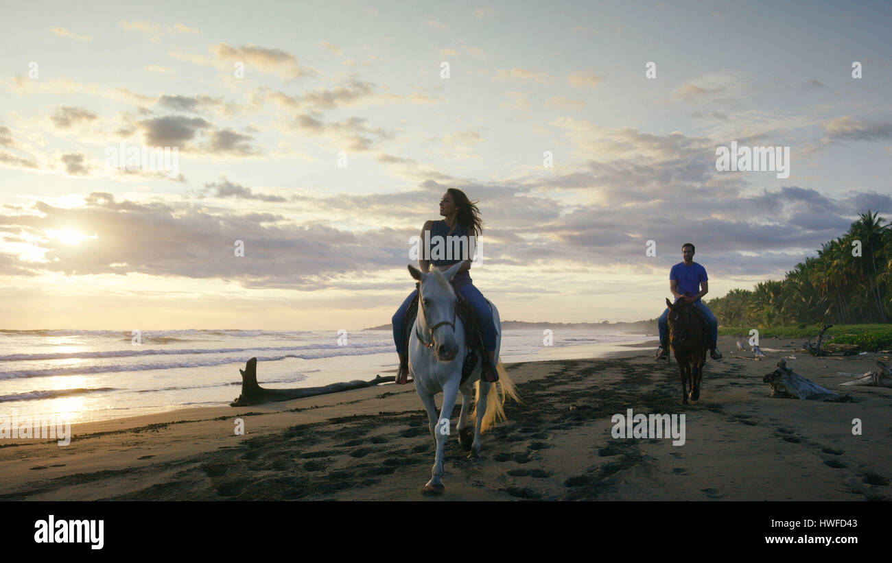 Couple de l'équitation sur la plage sur l'océan sous Ciel de coucher du soleil Banque D'Images