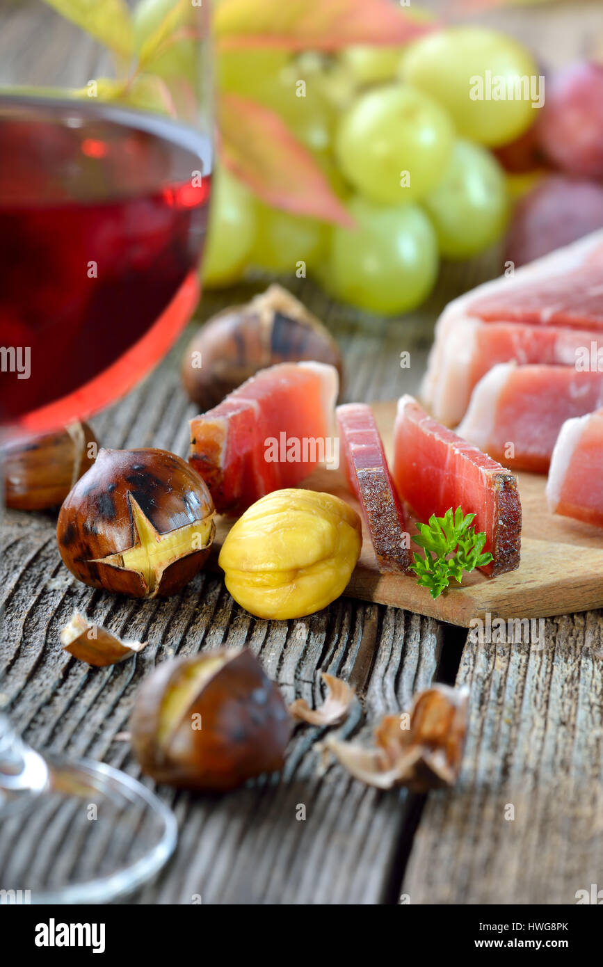 Châtaignes grillées avec du bacon du Tyrol du Sud et d'un verre de vin rouge sur une vieille table en bois, les raisins de l'arrière-plan Banque D'Images