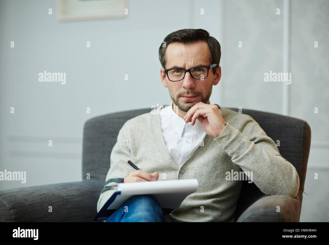 Portrait of handsome middle-aged psychologue en lunettes looking at camera, assis sur un fauteuil confortable et en remplissant la carte médicale Banque D'Images