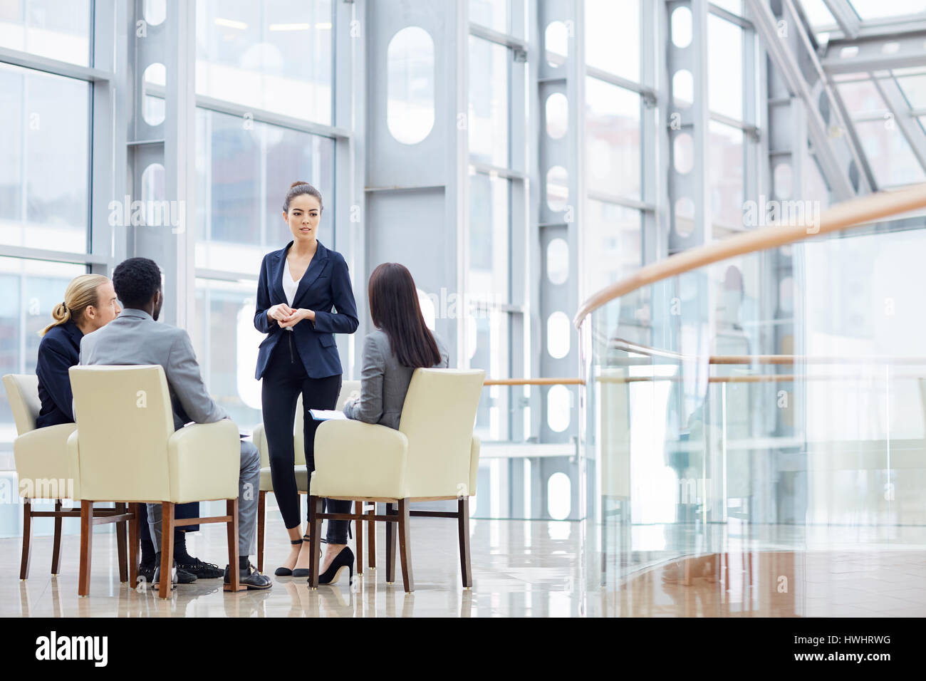 Groupe de quatre hommes d'affaires assis dans les fauteuils au cours de réunion, une femme giving presentation in glass hall d'immeuble de bureaux modernes Banque D'Images