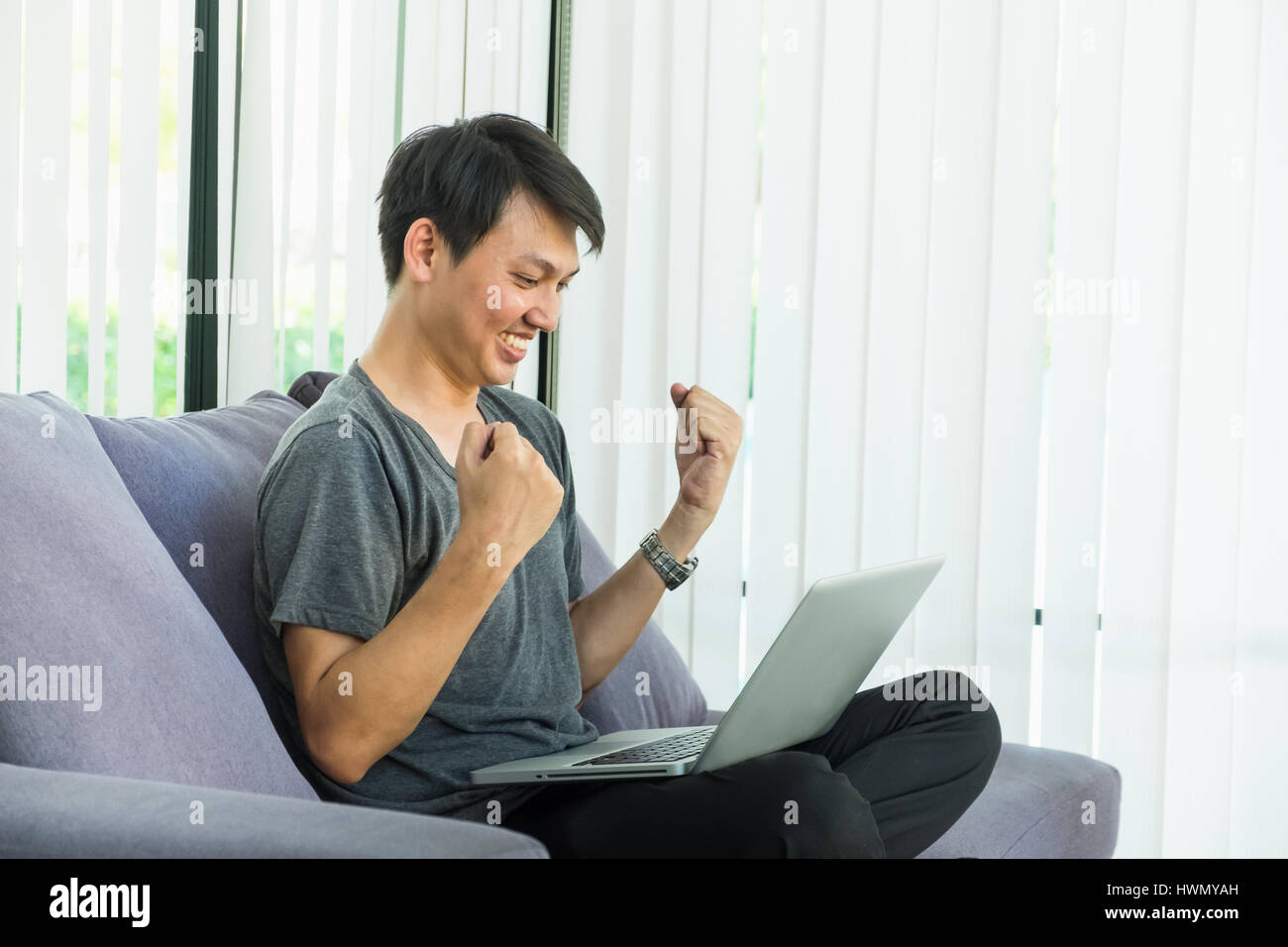 Jeune homme assis sur le canapé avec sentiment heureux jusqu'à bras posture lorsque vous regardez l'écran de l'ordinateur portable,succès célébration concept. Banque D'Images
