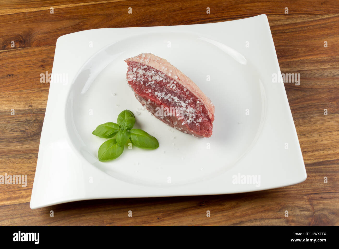 Tranche de viande de vache cru salé (picanha) et les feuilles de basilic sur un plat blanc et table en bois naturel d'époque - gros sel était utilisé Banque D'Images