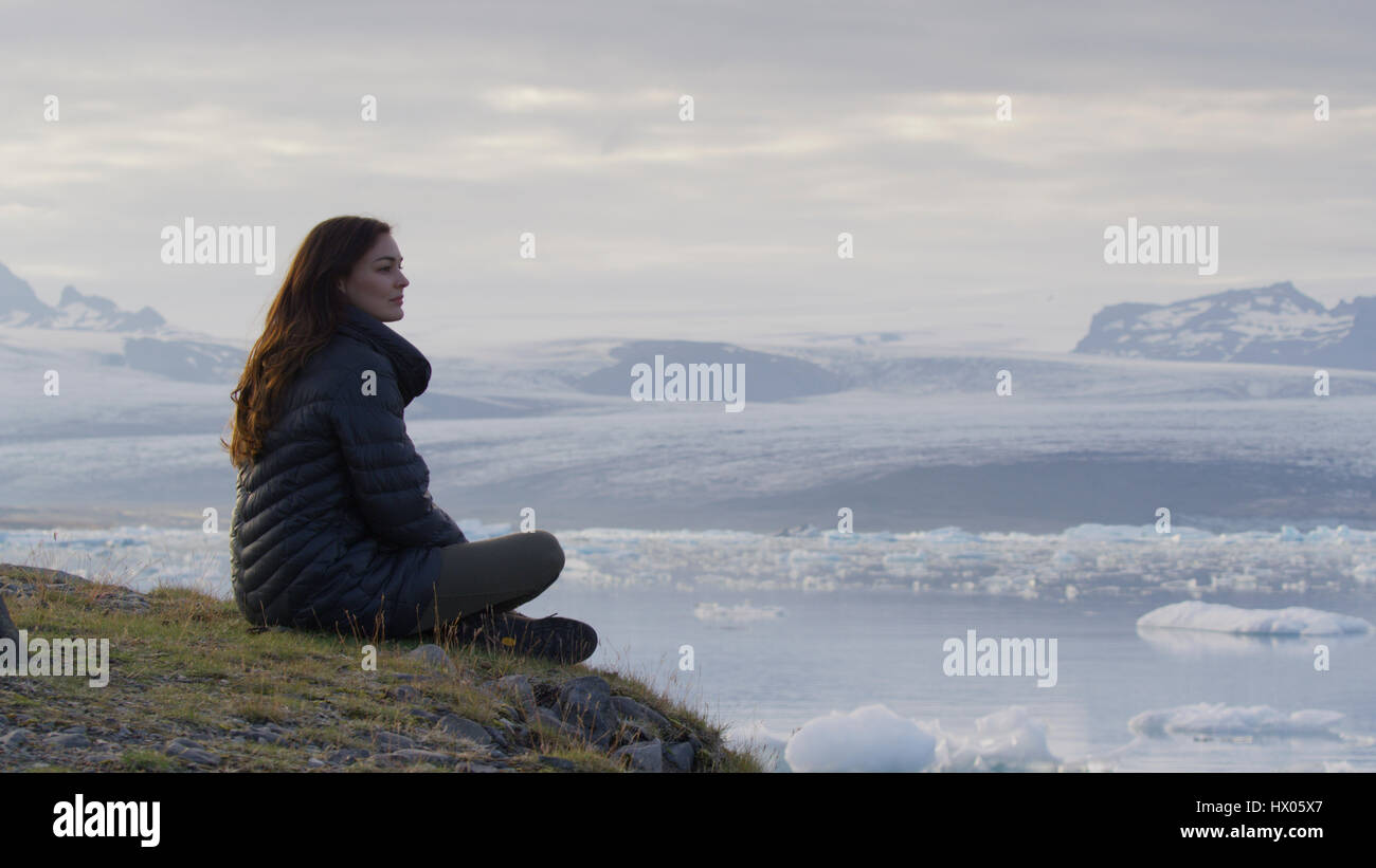 Vue de profil de femme assise sur l'herbe d'une colline donne sur le paysage et les glaciers gelés dans l'océan encore à distance sous ciel nuageux Banque D'Images