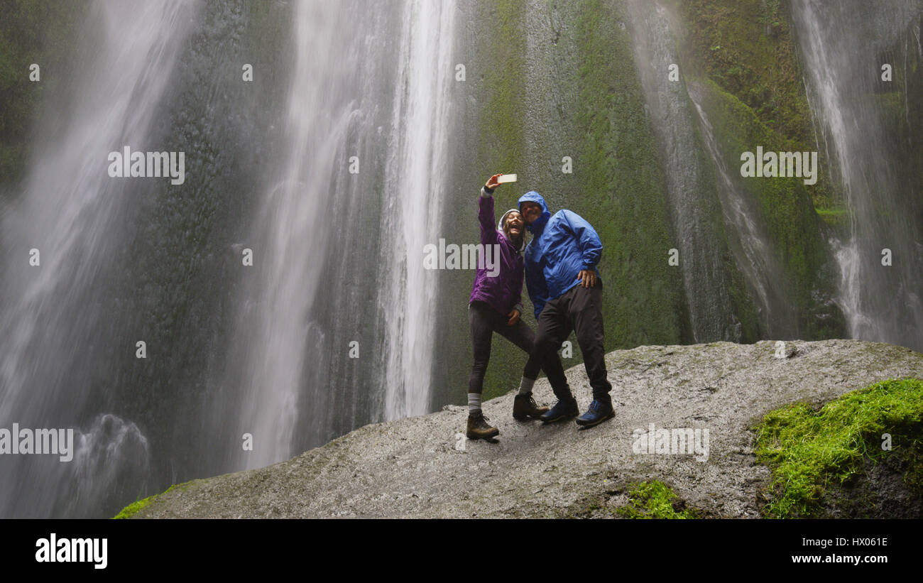 Low angle view of couple en tenant debout sur selfies smartphone boulder moussus près de cascade dans le paysage à distance Banque D'Images