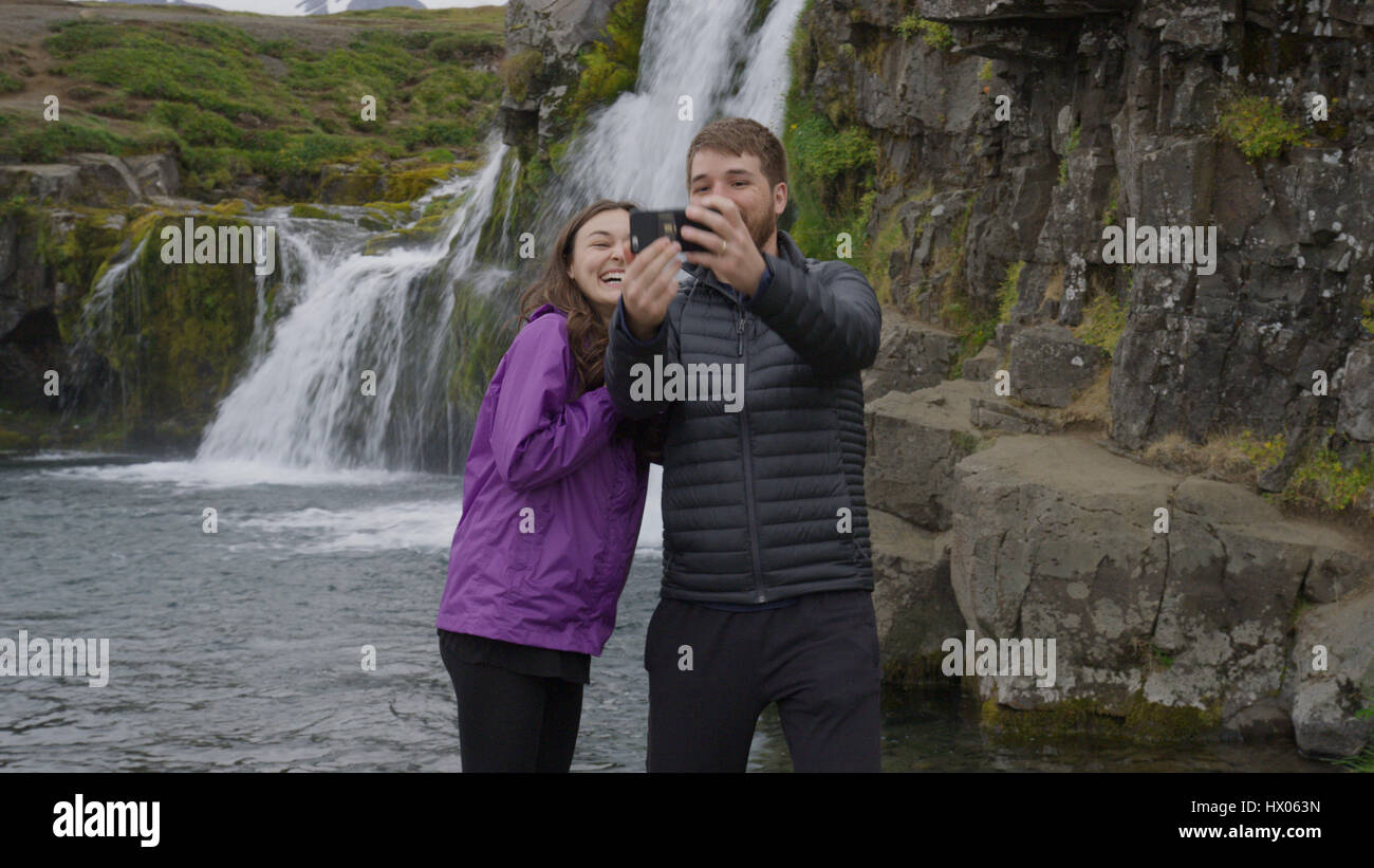 Petit ami et petite amie posant et en tenant près de waterfall selfies smartphone à distance sur les falaises de roche Banque D'Images