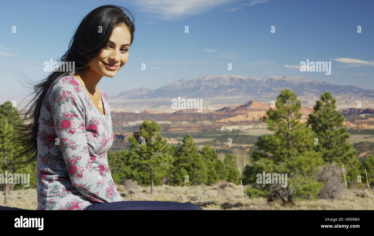 Portrait of smiling woman sitting près d'une vue panoramique sur les montagnes, arbres et broussailles de paysage à distance sous ciel bleu Banque D'Images