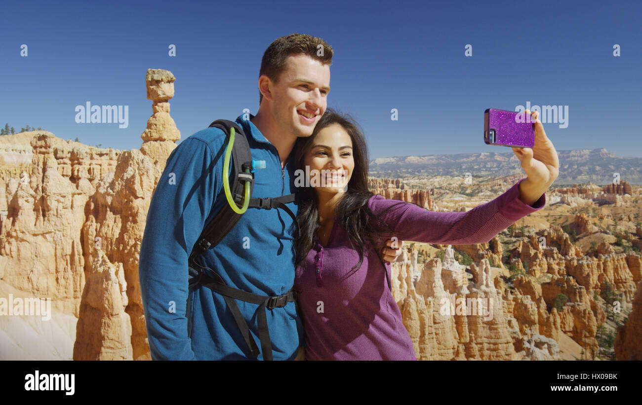 Petit ami et petite amie en tenant avec smartphone selfies près de formations rocheuses pittoresques dans le paysage à distance sous ciel bleu clair Banque D'Images