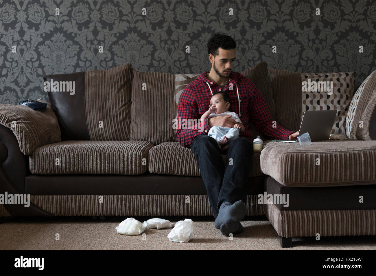 Jeune père, assis sur son canapé à la maison avec son petit fils sur son genou. Il travaille sur un ordinateur portable et il y a des couches sales sur le sol. Banque D'Images