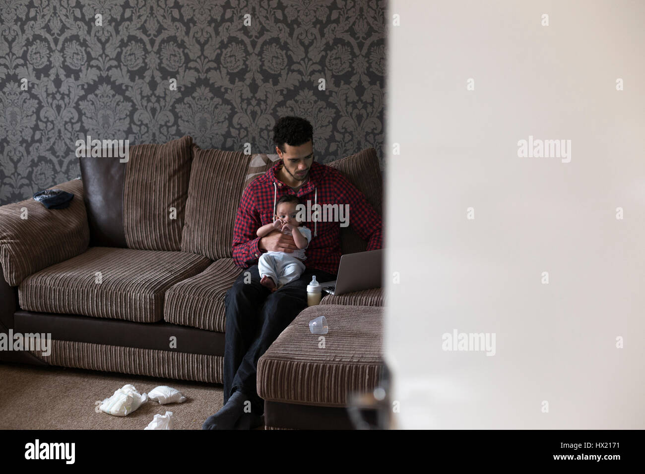 Jeune père, assis sur son canapé à la maison avec son petit fils sur son genou. Il travaille sur un ordinateur portable et il y a des couches sales sur le sol. Banque D'Images