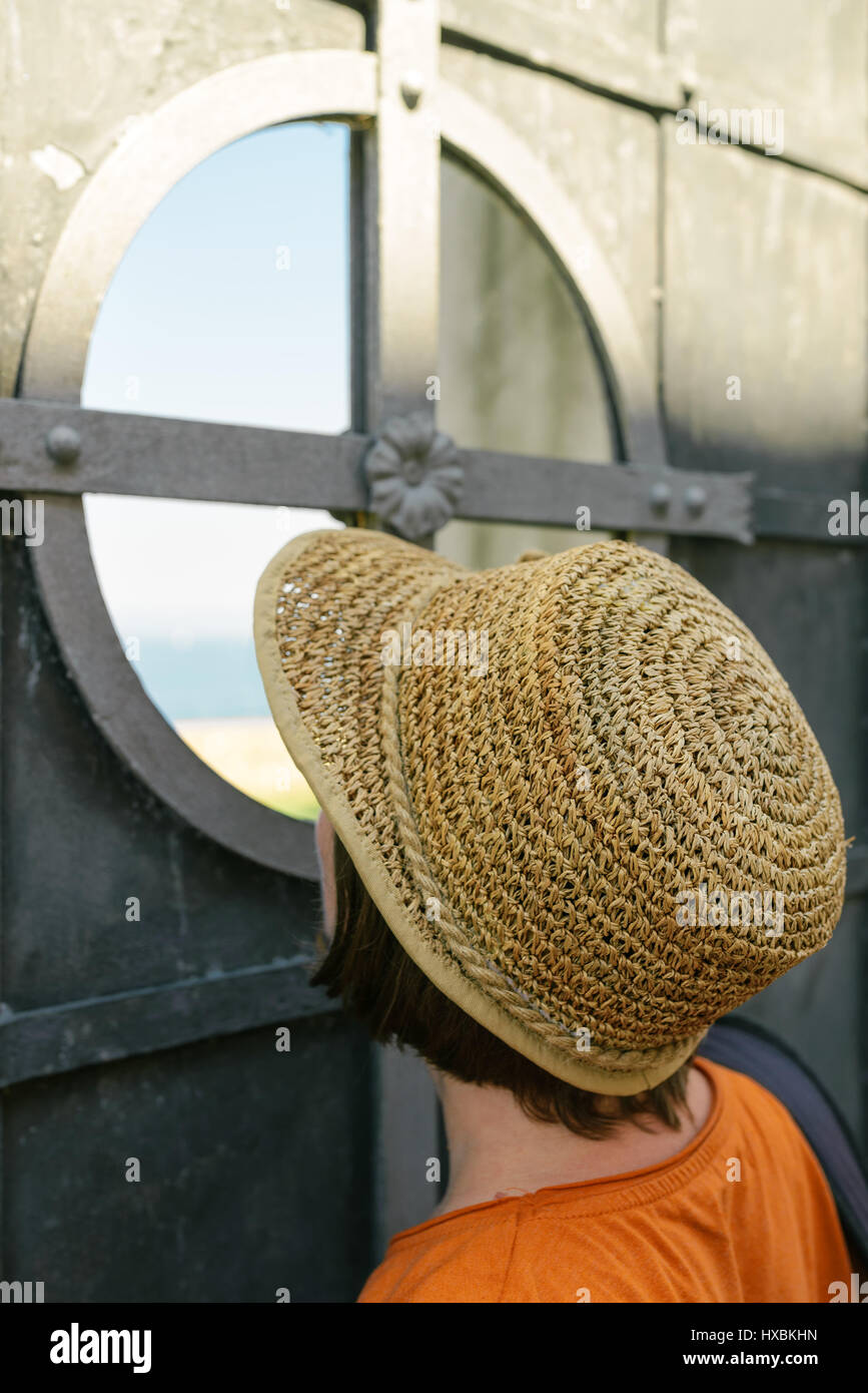 Curieux woman with hat looking through metal gate Banque D'Images