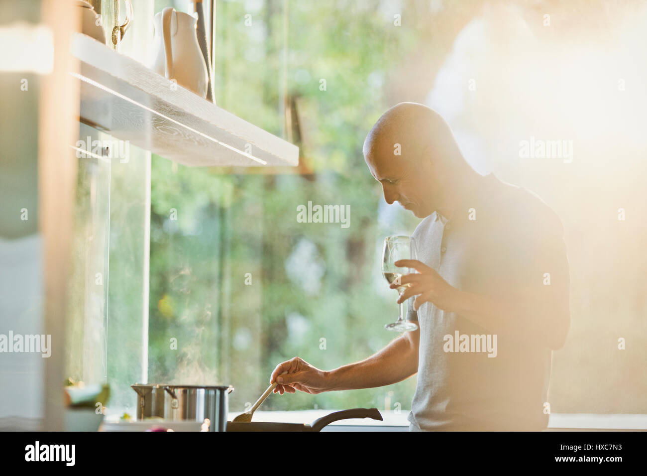 Mature man drinking white wine et cuisiner à la poêle dans cuisine ensoleillée Banque D'Images