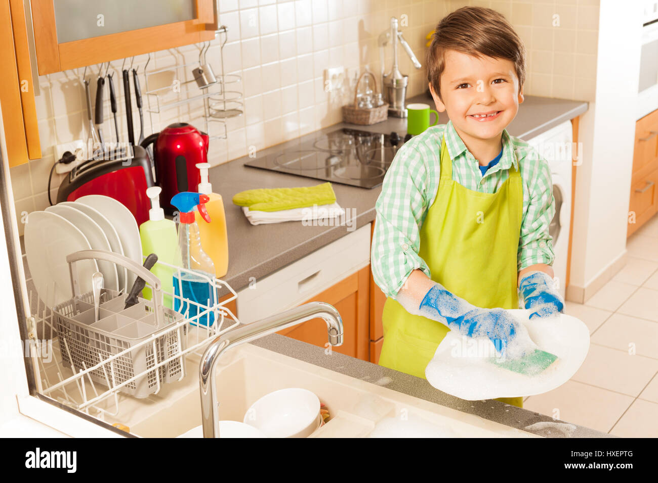 Peu jeune petit garçon la vaisselle dans la cuisine avec rdp et savon portant un tablier smiling to camera Banque D'Images