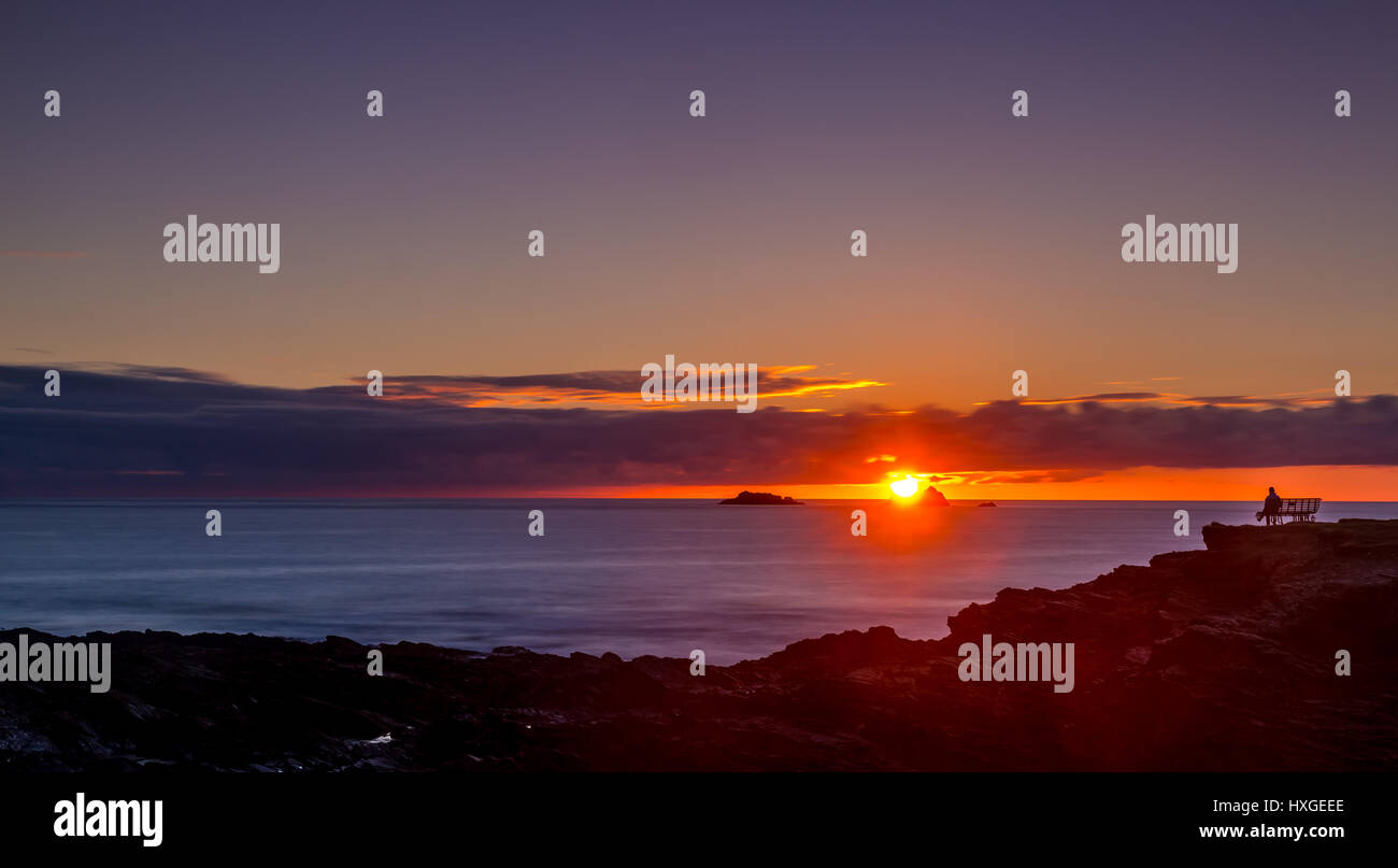 Personne assis sur banc coastside donnant sur le magnifique coucher de soleil sur l'horizon. La baie de Treyarnon, Cornwall. Banque D'Images