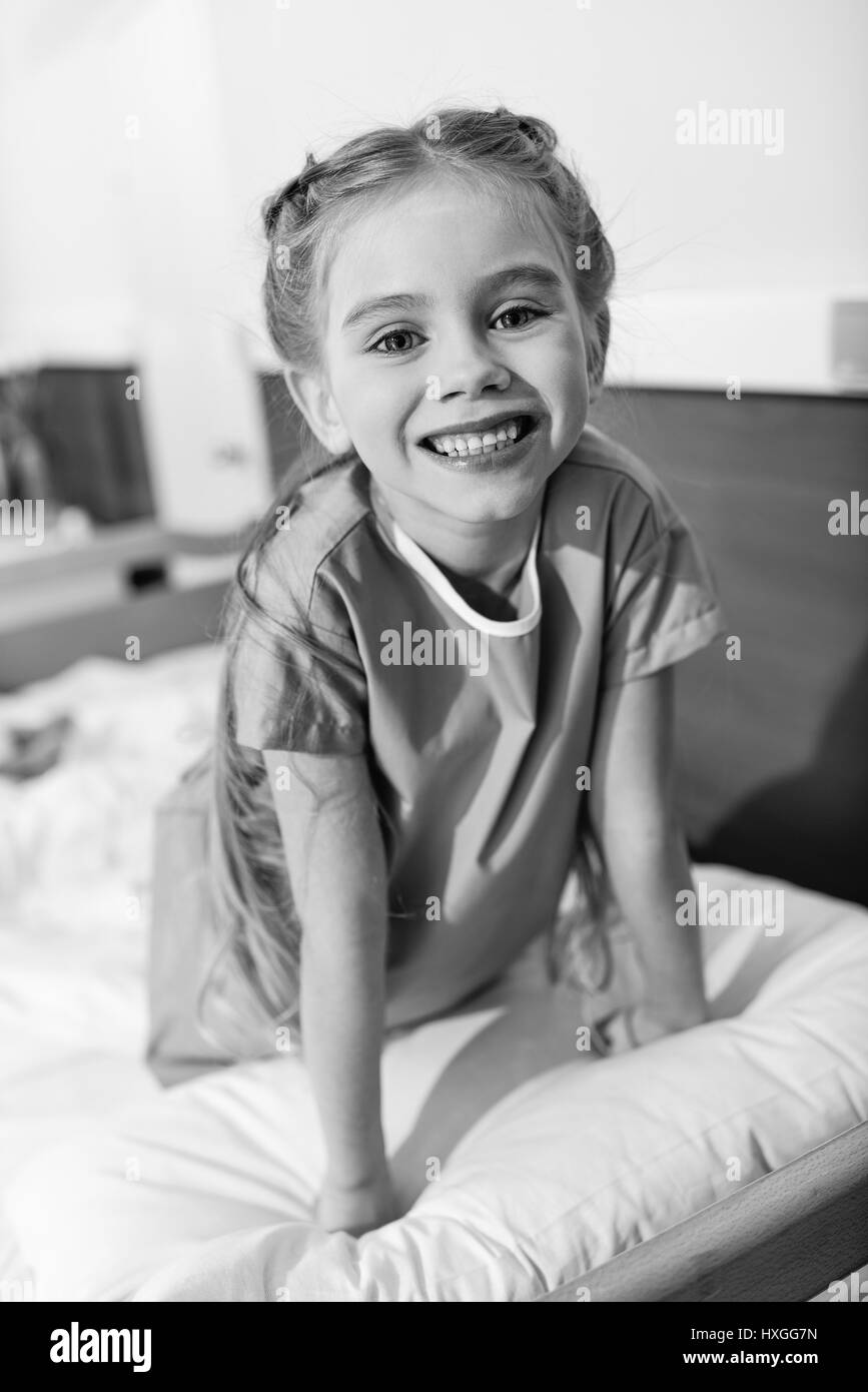 Cute little girl sitting on hospital bed and smiling at camera, photo en noir et blanc Banque D'Images