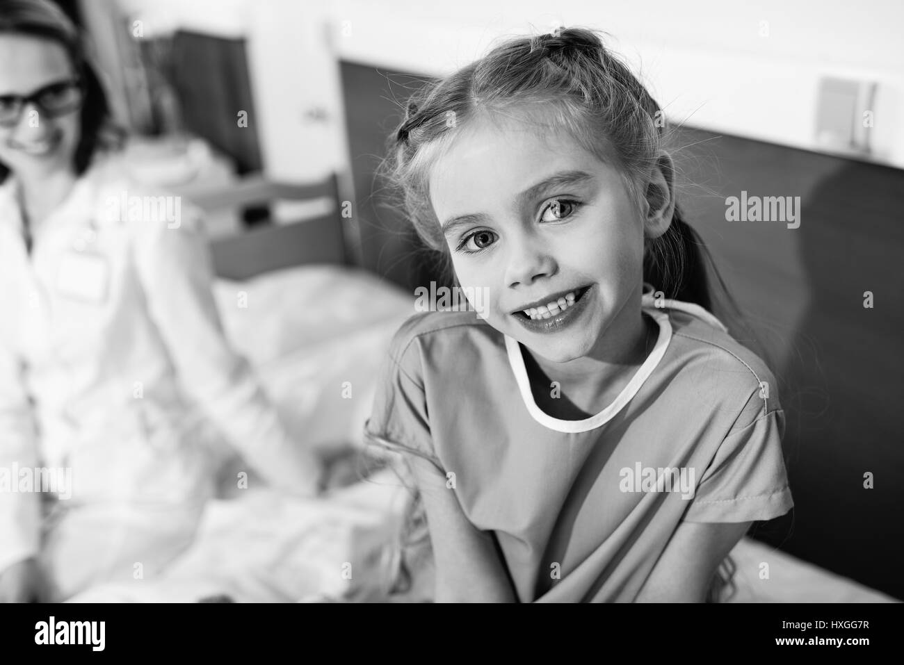 Cute little girl sitting on hospital bed and smiling at camera, photo en noir et blanc Banque D'Images
