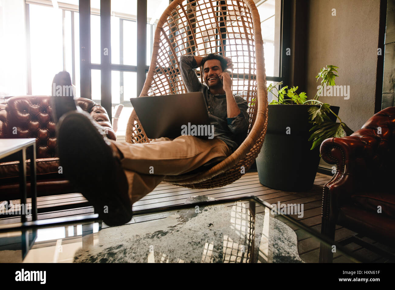 Jeune homme d'affaires assis sur un fauteuil suspendu en rotin dans le salon bureau avec ordinateur portable et talking on mobile phone. Male executive office Banque D'Images