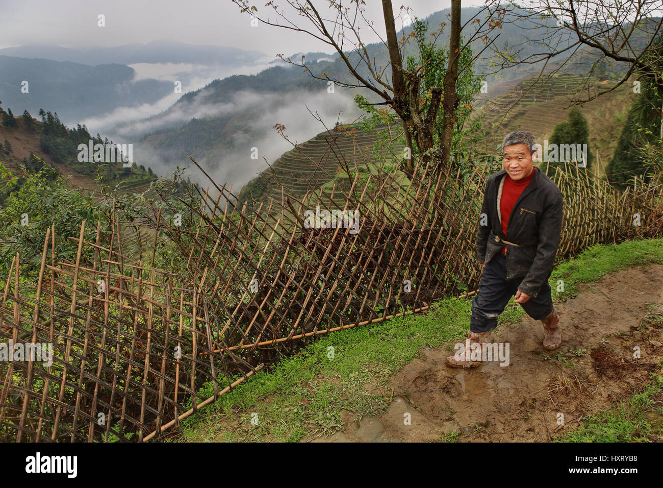 Village Pingan, Province du Guangxi, Chine - 5 Avril 2010 : Asian man, portant des bottes en caoutchouc, la marche sur route de boue, au-delà de la barrière sur l'arrière-plan Banque D'Images