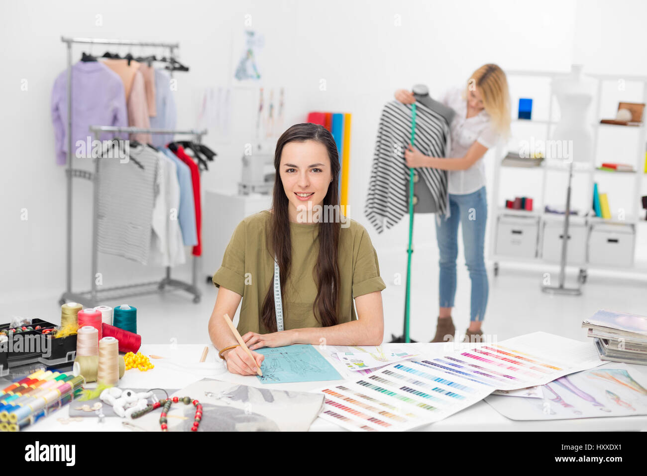 Deux jeunes stylistes collaborer pour de nouveaux vêtements collection dans leur atelier , l'un en premier plan assis au bureau et l'autre sur dessin Banque D'Images