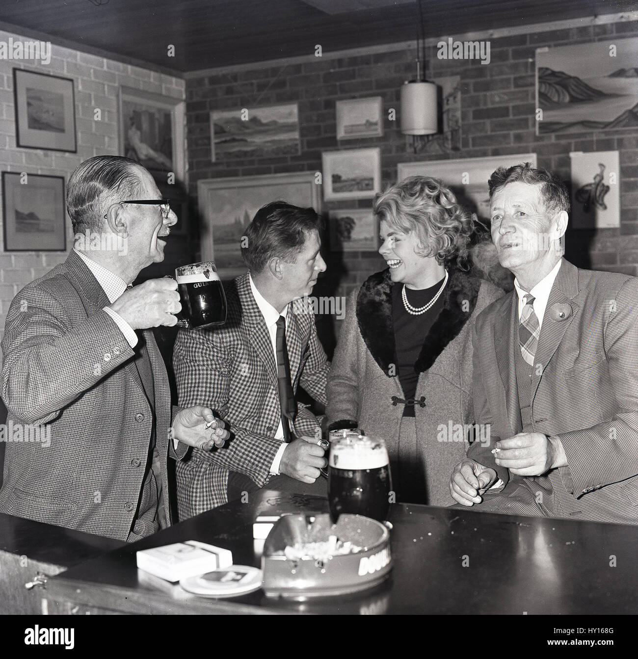Années 1960, historiques, glamour femme dans un manteau avec un revers de fourrure dans un bar avec des collègues masculins de déguster une boisson et l'allume après le travail, England, UK Banque D'Images
