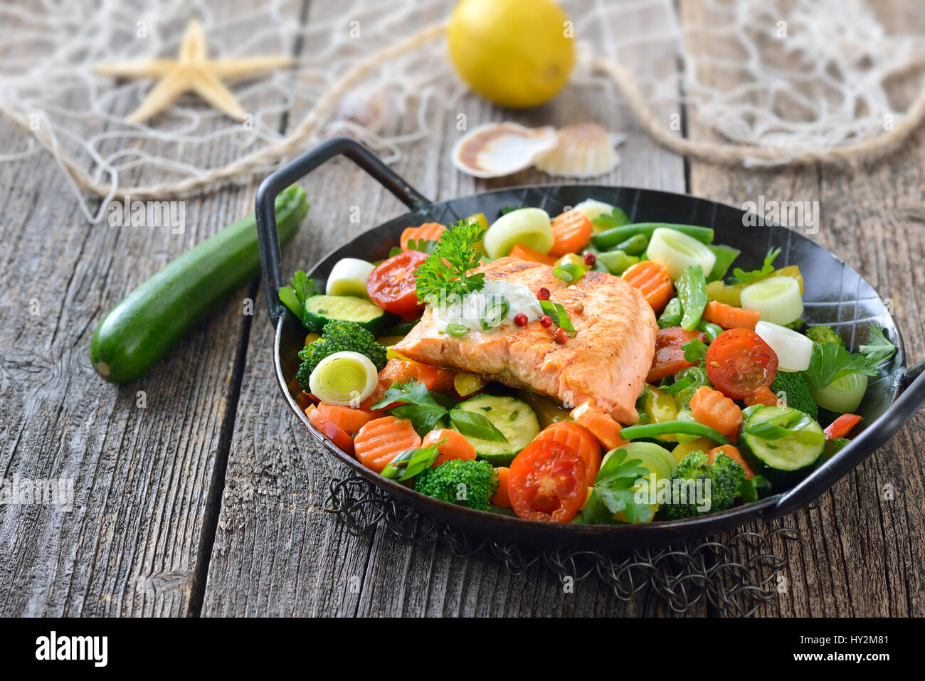 Filet de saumon poêlé sur des légumes colorés servi dans une poêle, un citron et un filet de pêche avec une étoile de mer et coquillages en arrière-plan Banque D'Images