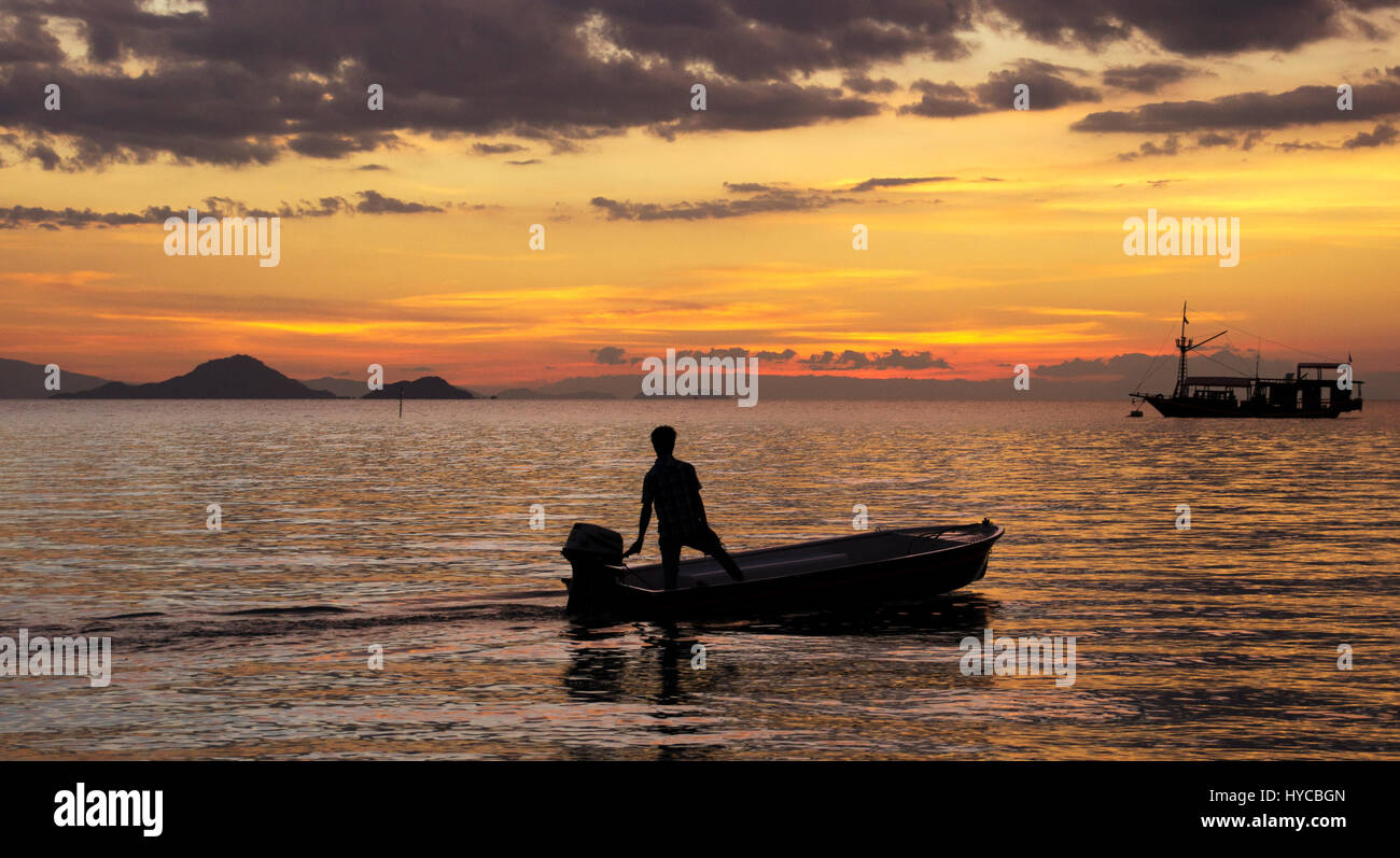 Silhouette d'un pêcheur sur un petit bateau de pêche avec moteur hors bord sur l'océan à la tombée de la nuit avec un coucher du soleil orange et un autre bateau à la zone Banque D'Images