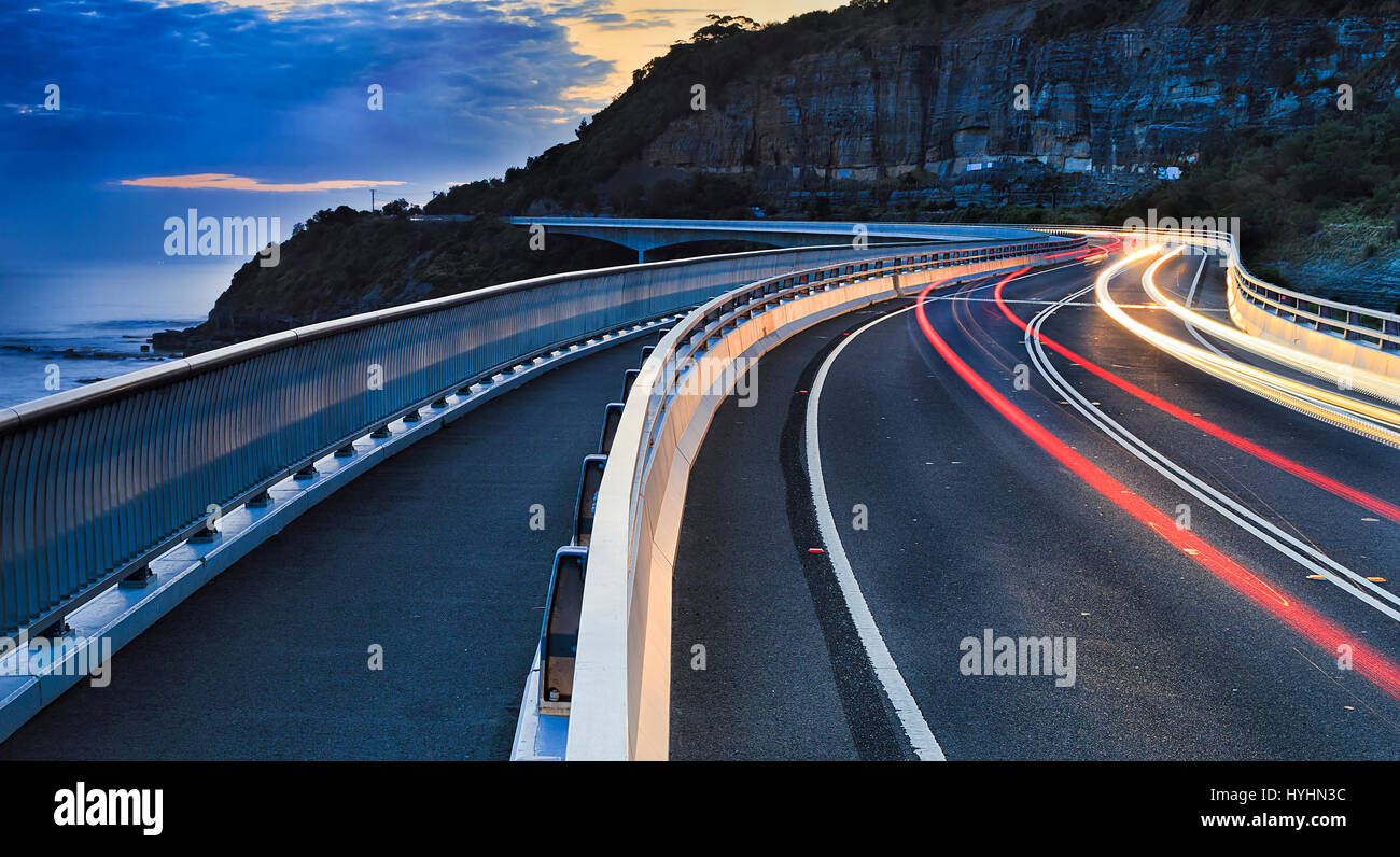 Phares des voitures floues déménagement sur Sea Cliff bridge - partie de Grand Pacific Drive dans le NSW, Australie, au coucher du soleil. Banque D'Images