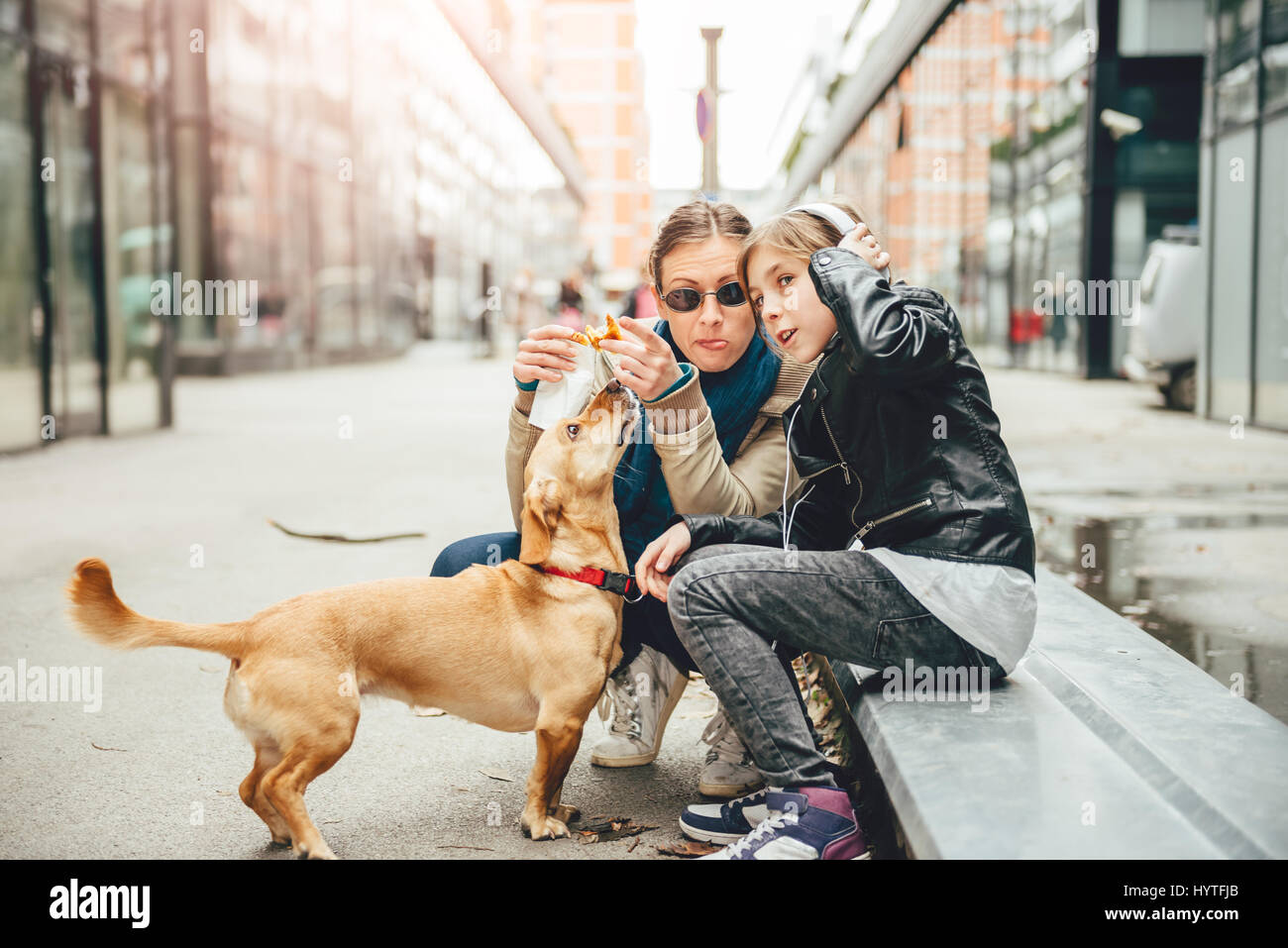 Mère eating sandwich et flatter un chien bien que fille d'écouter de la musique dans la rue Banque D'Images