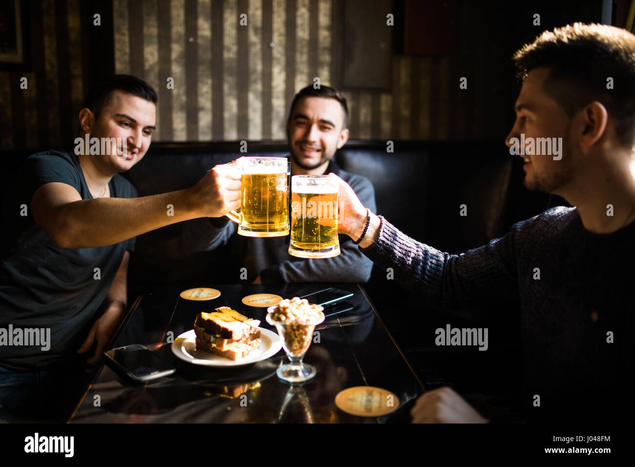 Trois jeunes hommes sont souriants et qui retentit verres de bière ensemble tout en restant assis dans un pub Banque D'Images