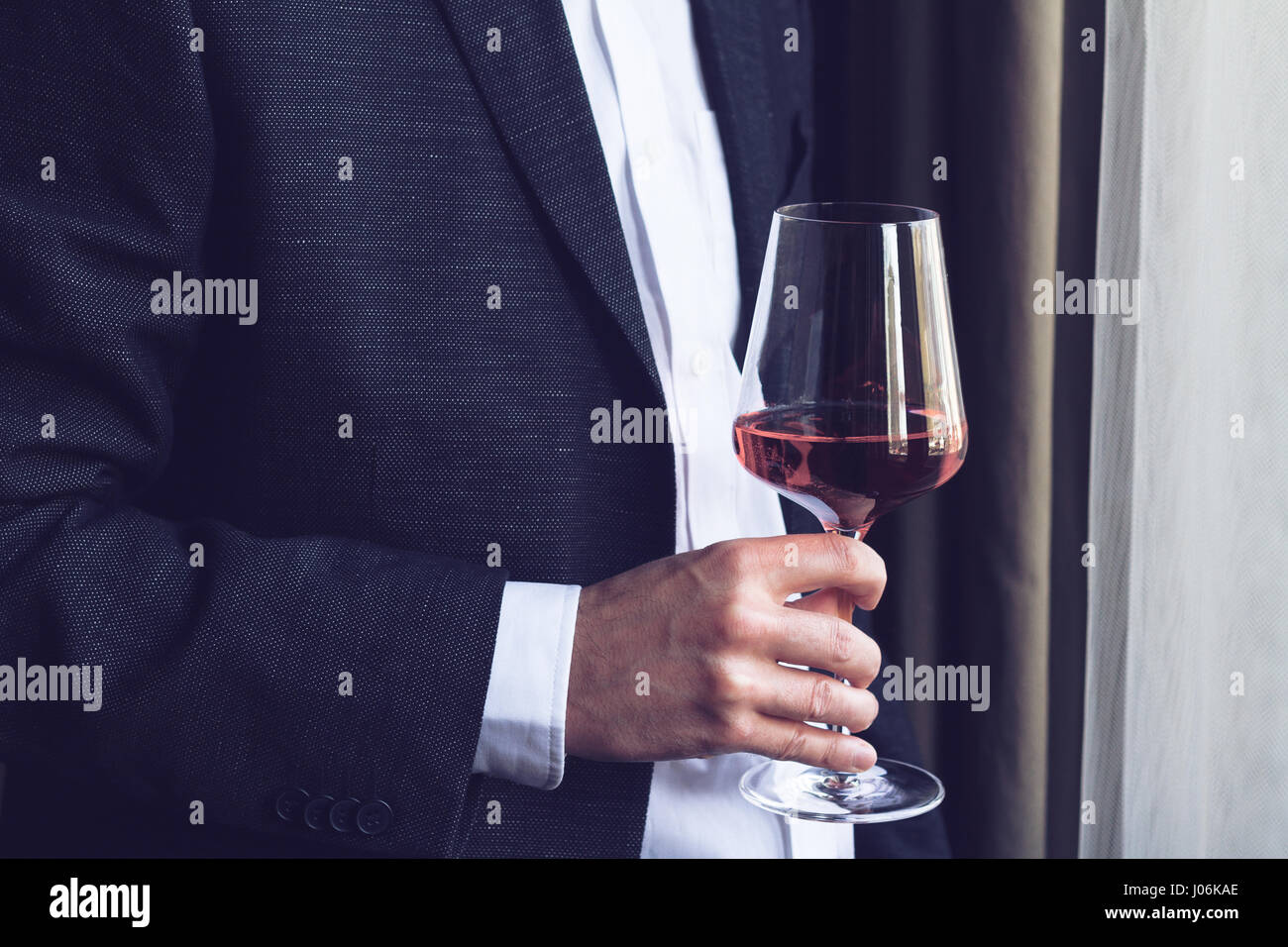 Close up of Horizontal Caucasian man en costume noir et chemise blanche tenant un grand verre à vin rose lors d'un événement par la fenêtre d'éclairage naturel Banque D'Images