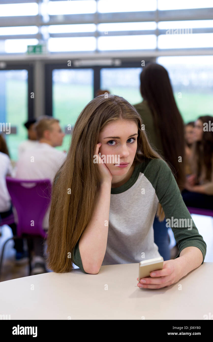Étudiante assise sur son propre à l'école avec un smartphone. Elle regarde la caméra avec une expression triste. Banque D'Images