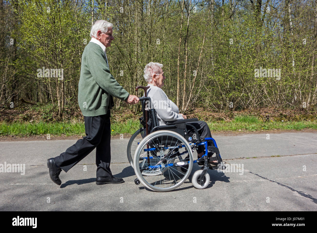 Mari à la retraite âgées handicapées prendre épouse dans fauteuil roulant pour une promenade dans le parc lors d'une journée ensoleillée au printemps Banque D'Images