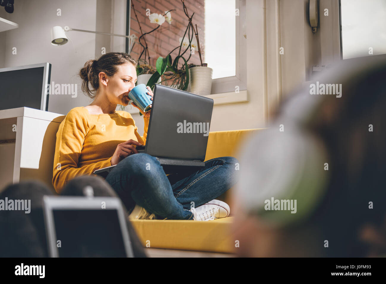 Femme portant le chandail jaune jaune assis sur un canapé à boire du café et à l'aide d'ordinateur portable à la maison Banque D'Images Femme portant le chandail jaune jaune assis sur un canapé à boire du café et à l'aide d'ordinateur portable à la maison Banque D'Images