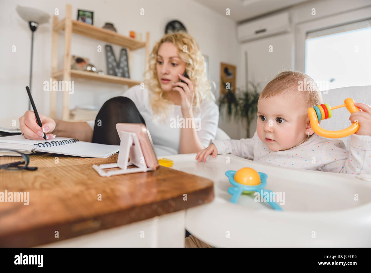 Regarder bébé excité alors que smart phone mother talking on smart phone et l'écriture des notes au bureau à domicile Banque D'Images