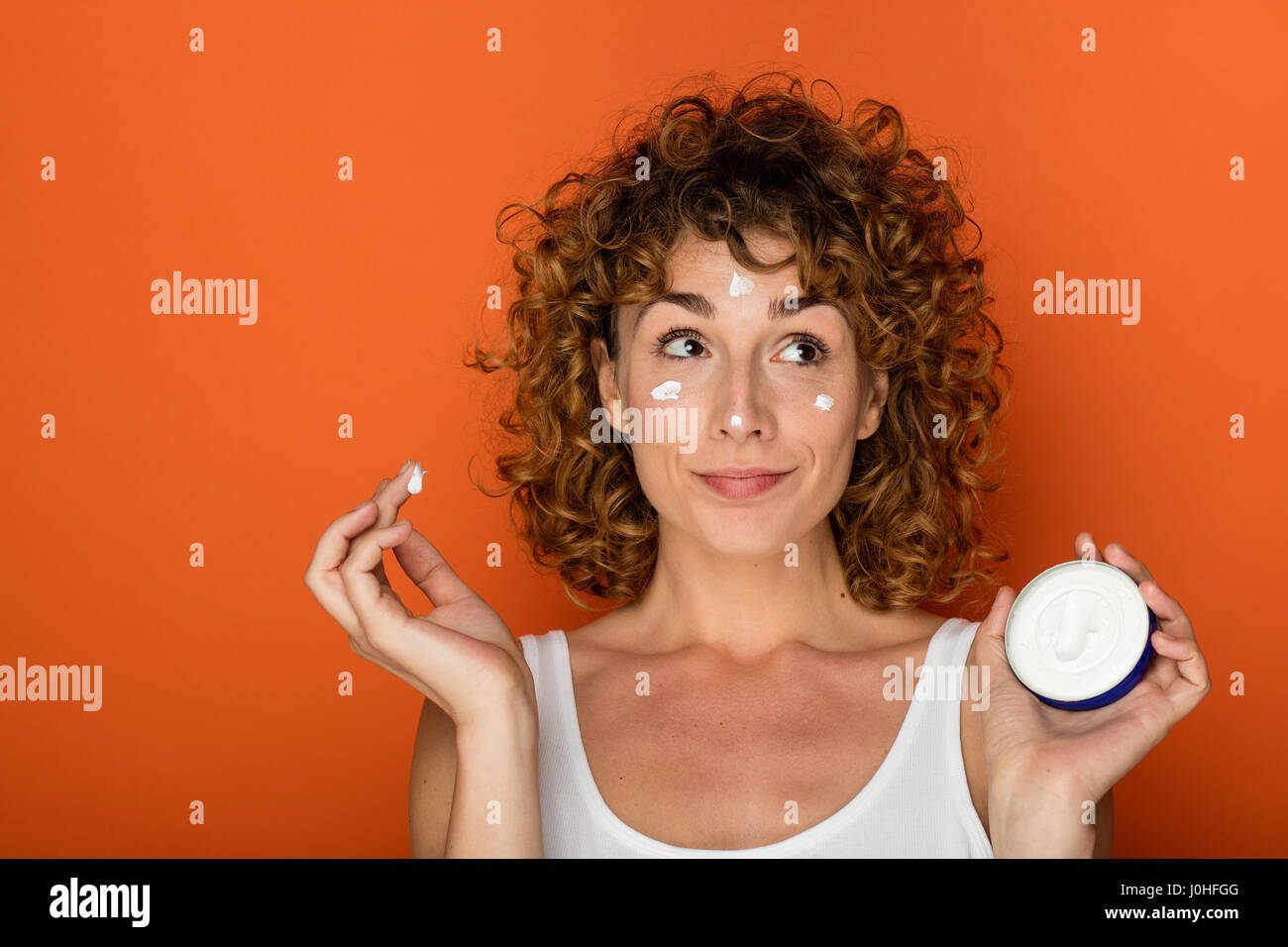 Young woman holding bouclés et à l'aide d'une petite boîte de crème de peau sur fond orange Banque D'Images