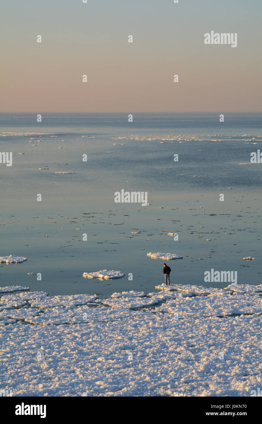 Un homme se tient sur le bord d'une plaque de glace et regarde vers le bas dans l'eau. L'immensité du lac Michigan s'étend au-delà de lui. Banque D'Images