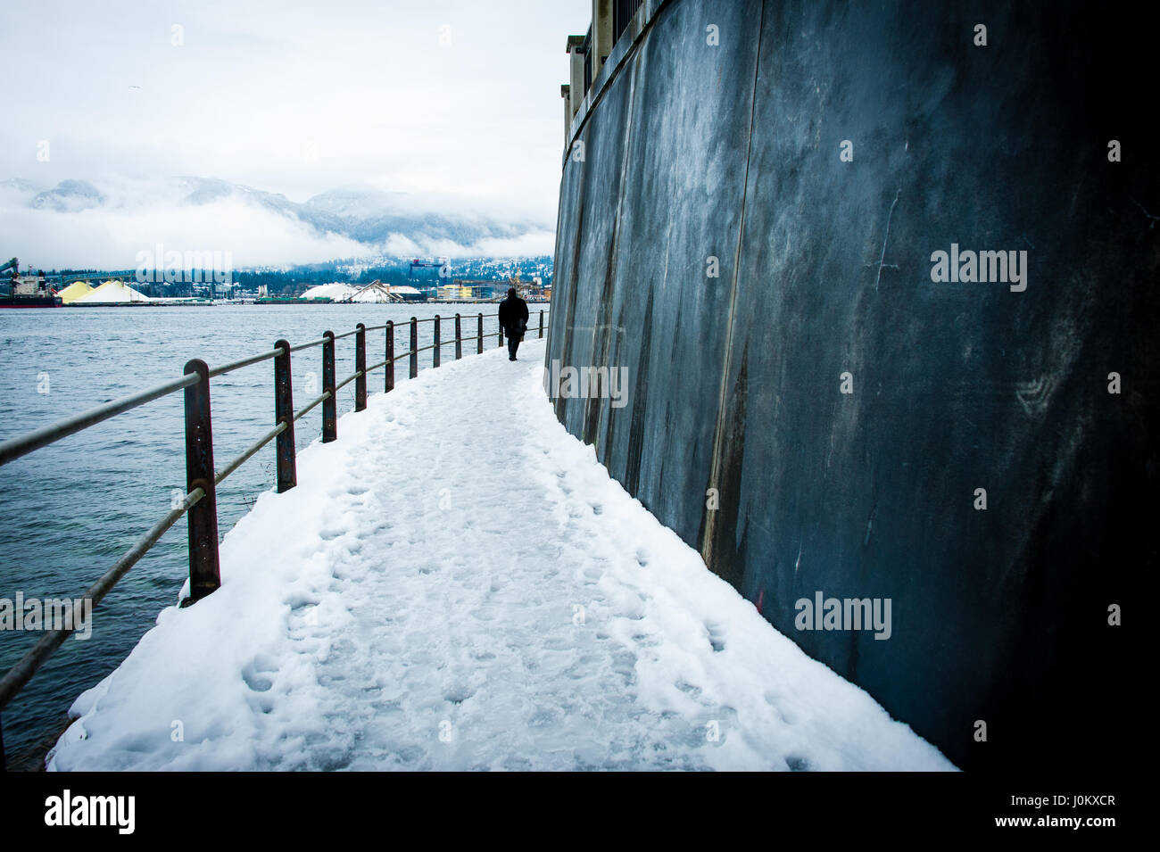 Seul l'homme en long manteau noir marcher le long d'un chemin couvert de neige avec clôture le long de l'océan. Grand mur sur le côté droit, site industriel en arrière-plan. Banque D'Images