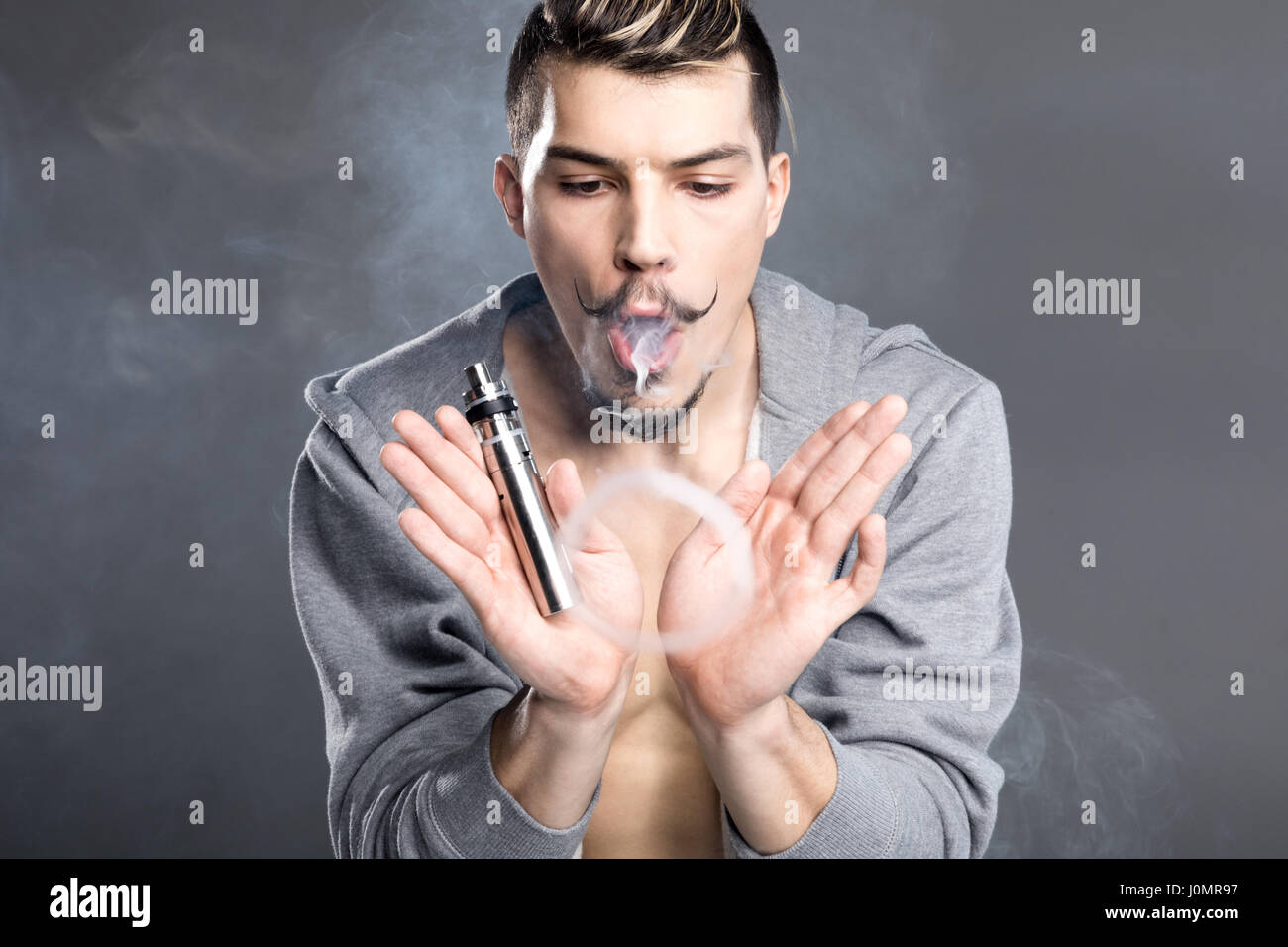 Jeune homme barbu tenant cigarette électronique et vaping sur gris Banque D'Images