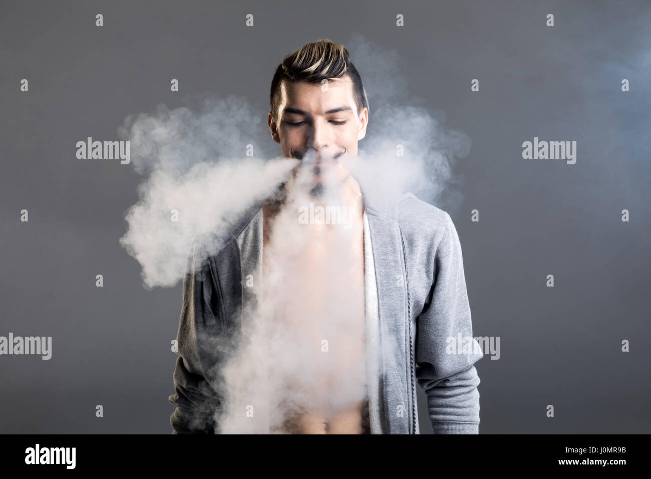 Jeune homme barbu souriant vaping et en soufflant la fumée sur gray Banque D'Images