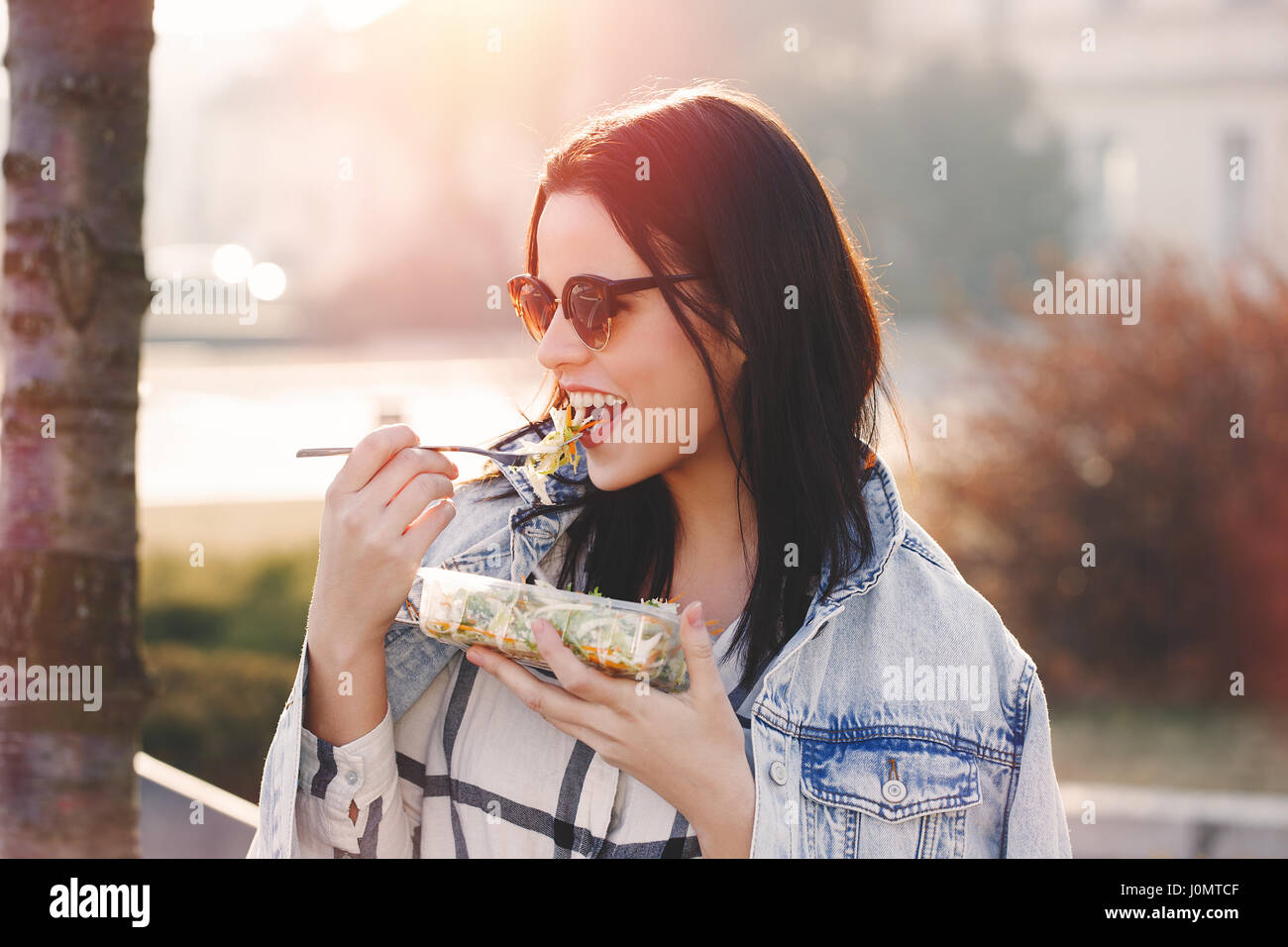 Jeune femme brune à lunettes de manger une salade fraîche au coucher du soleil en plein air, à l'écart Banque D'Images
