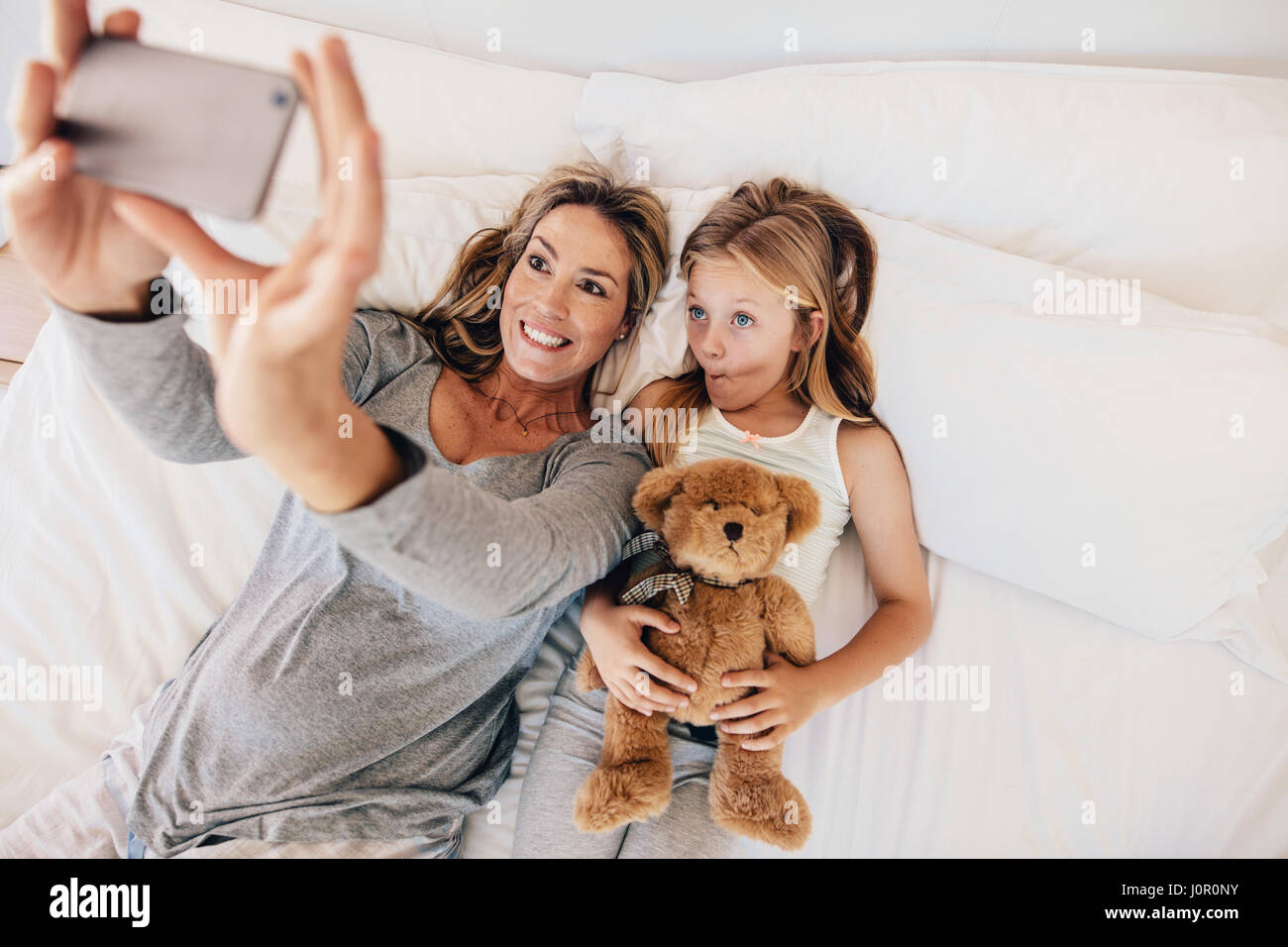 Mother and Daughter lying on bed and taking self portrait avec téléphone mobile. Femme tenant une petite fille avec des selfies holding ours au lit. Banque D'Images