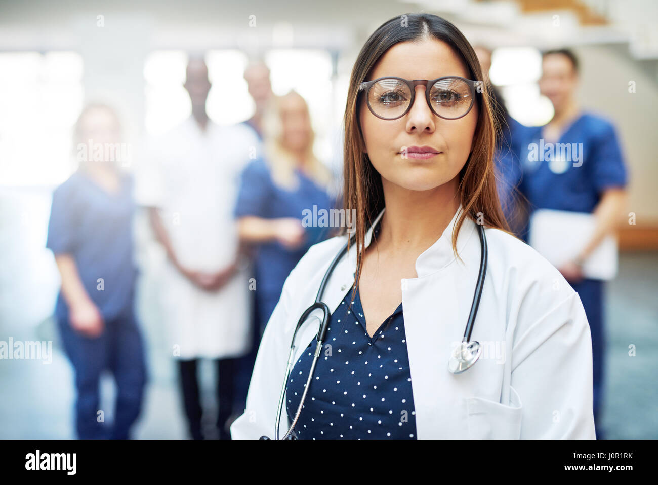 Jeune femme médecin confiant réfléchie à la caméra au comité permanent en clinique, en face de l'équipe médicale Banque D'Images