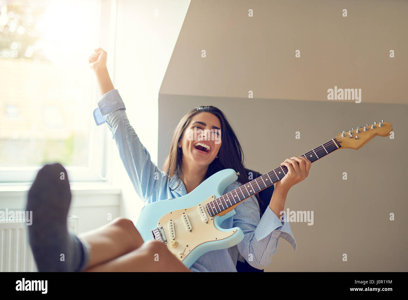Beau chant unique femme jouant de la guitare avec bras soulevé et les pieds sur la table dans la chambre avec fenêtre lumineuse Banque D'Images