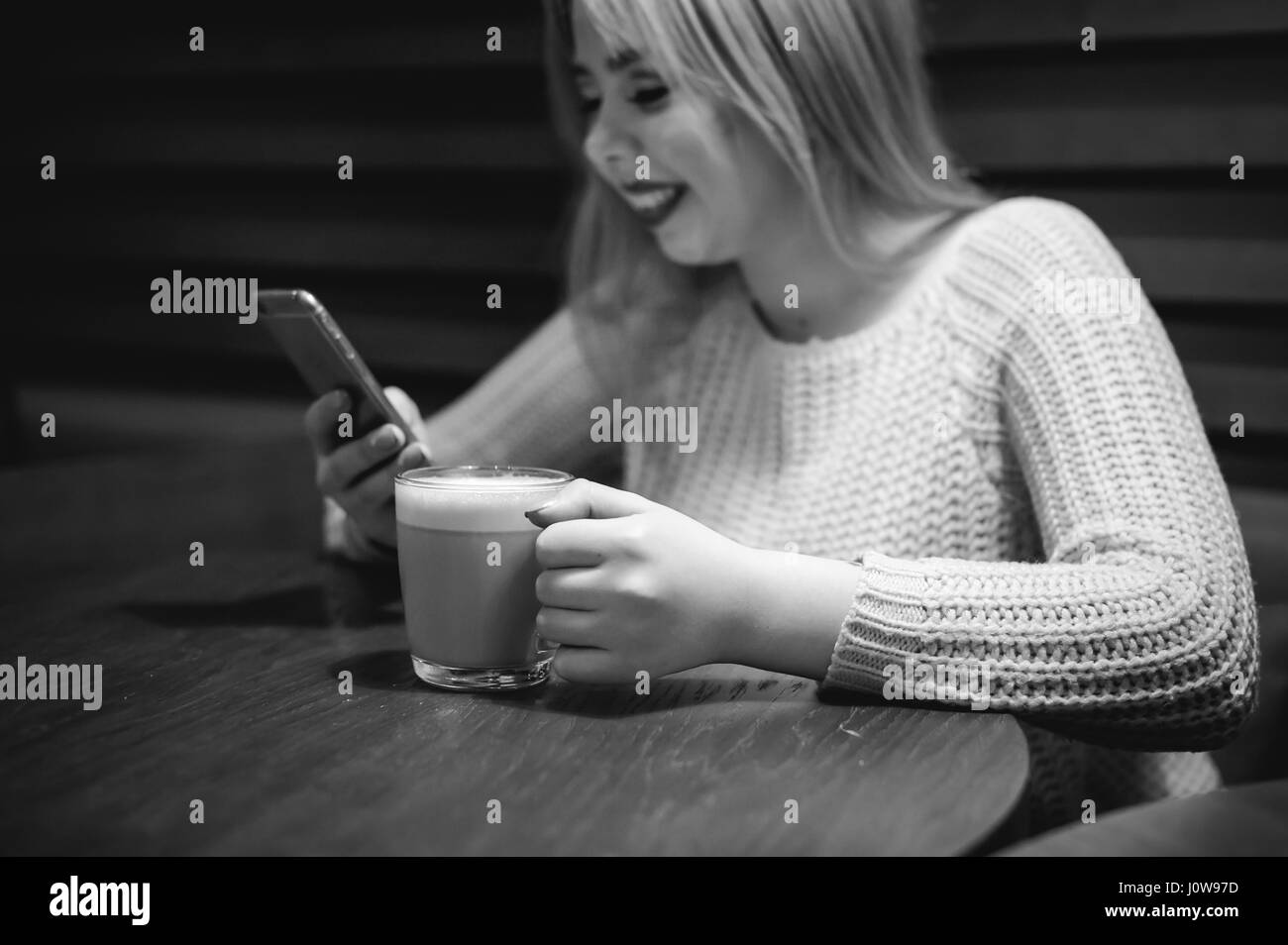 Jeune femme blonde dans un chandail tricoté assis à une table dans un café avec une tasse de café dans ses mains, en passant du temps seuls, faisant internet communicat Banque D'Images