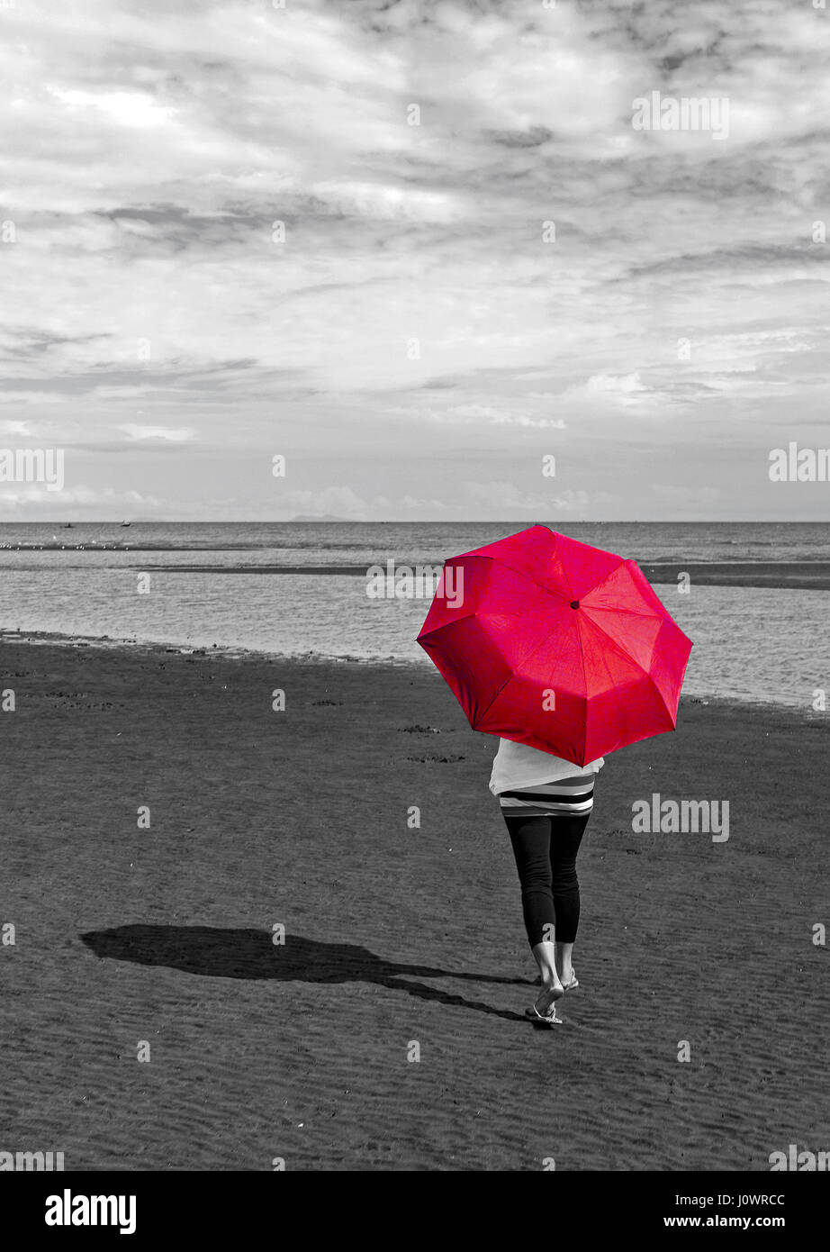 Une femme marche sur la plage avec un parapluie rouge. Cette photo est une goutte de couleur avec une conversion en niveaux de gris à l'exception du parapluie. Banque D'Images