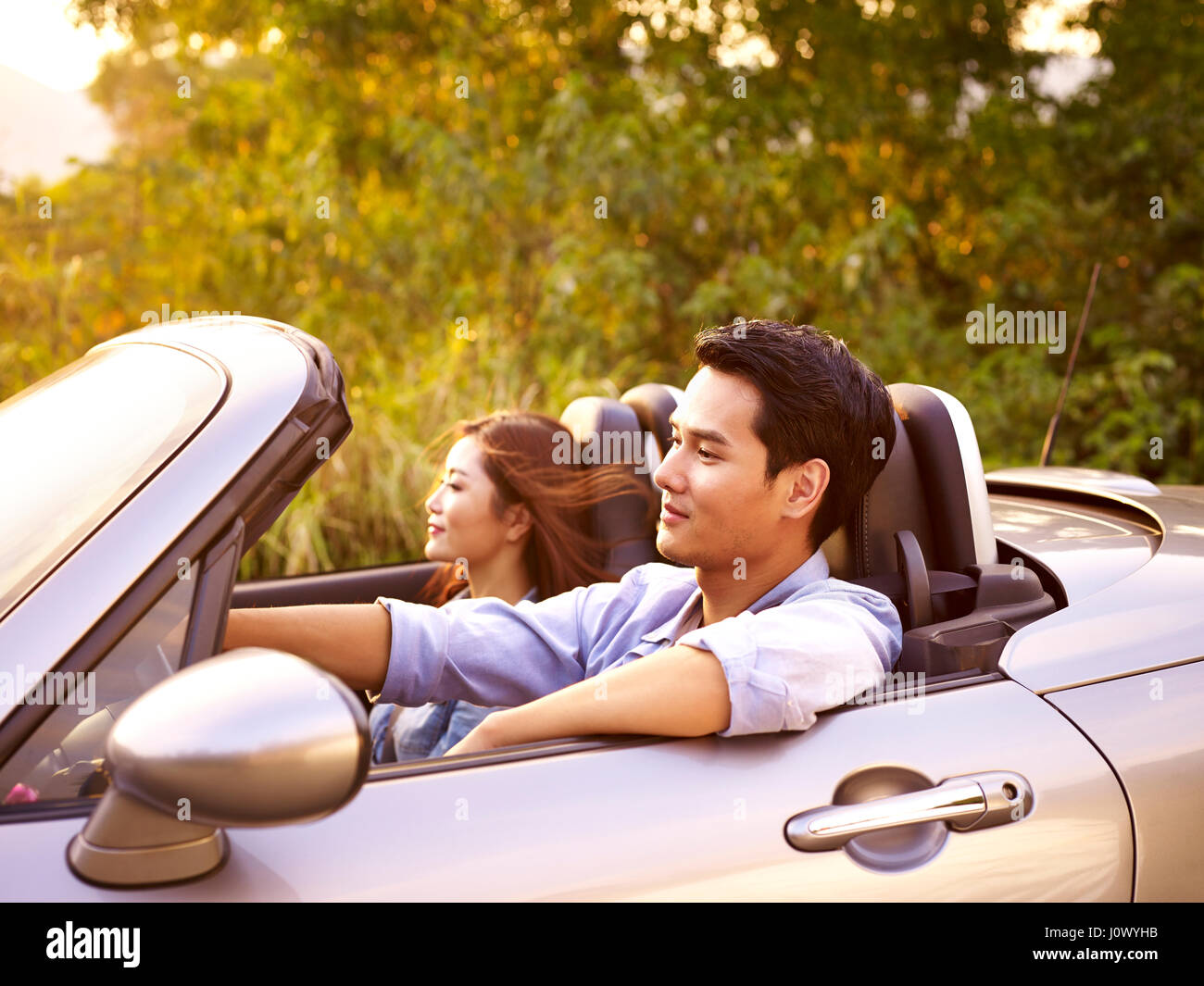 Young Asian couple riding dans un cabriolet sport car au coucher du soleil. Banque D'Images