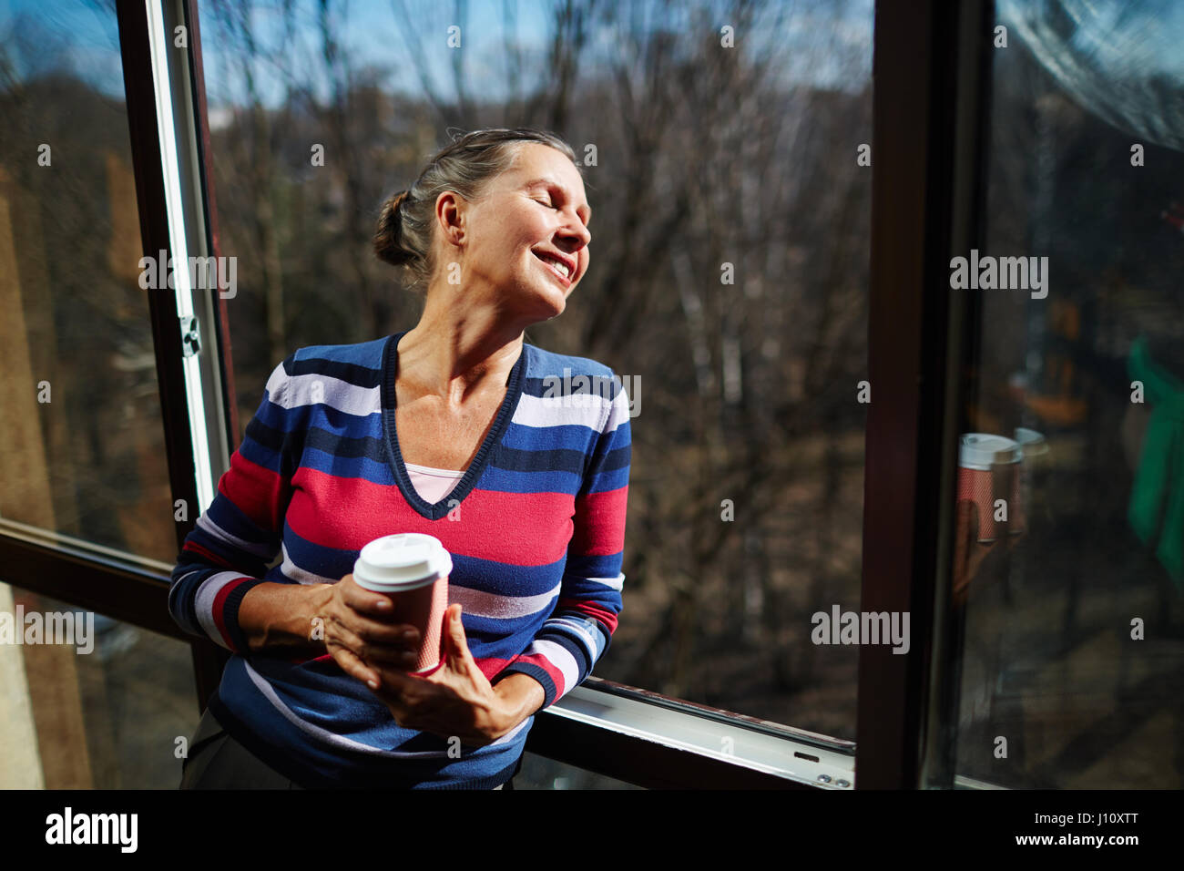 Heureux senior woman avec verre de boisson chaude s'appuyant sur la fenêtre ouverte Banque D'Images