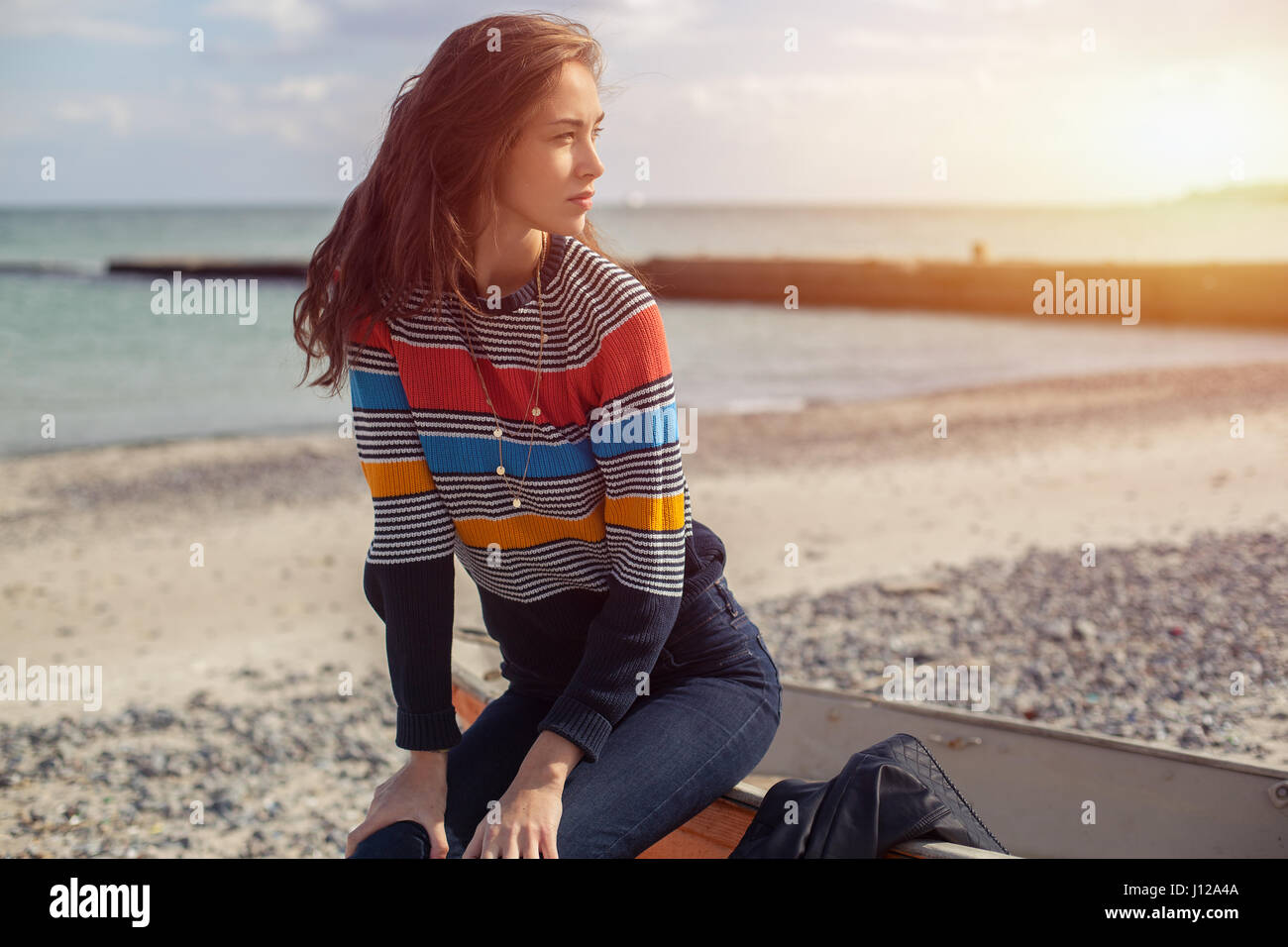 Une jeune fille assise sur le côté près d'un voile rouge sur la plage de la  mer avec magnifique coucher de soleil sur l'arrière-plan. Femme en jeans,  pull et gilet sur le