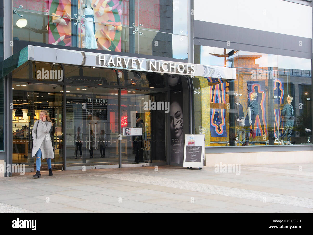 L'extérieur du magasin Harvey Nichols dans le centre de Manchester, Angleterre, avec la fenêtre affiche colorée et une femme de quitter le client boutique. Banque D'Images