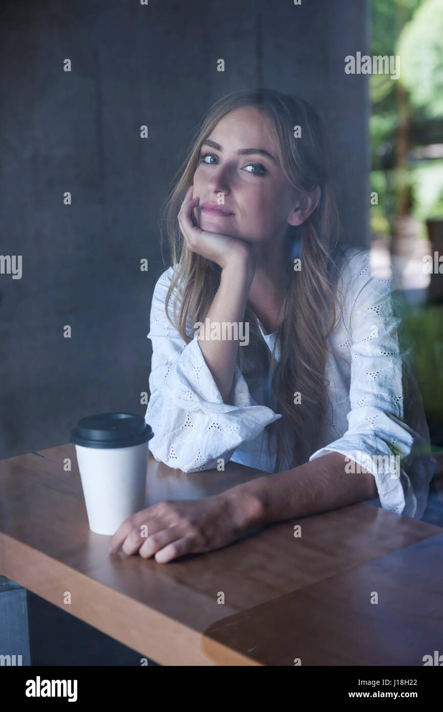 Jeune femme sérieuse assis dans un café avec café photographié par la fenêtre en verre de l'extérieur Banque D'Images