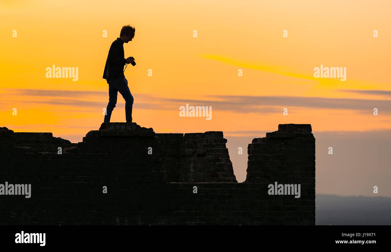 Tr8lhouette d'un photographe de marcher le long d'un mur dans la soirée, la tenue d'un appareil photo, avec un ciel orange à partir de la faible soleil du soir. Banque D'Images